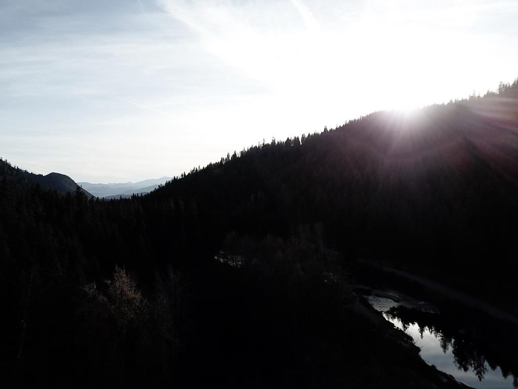 A silhouetted forested ridge backlit by the sun, with a river curving along the bottom right and distant mountains in the haze.
