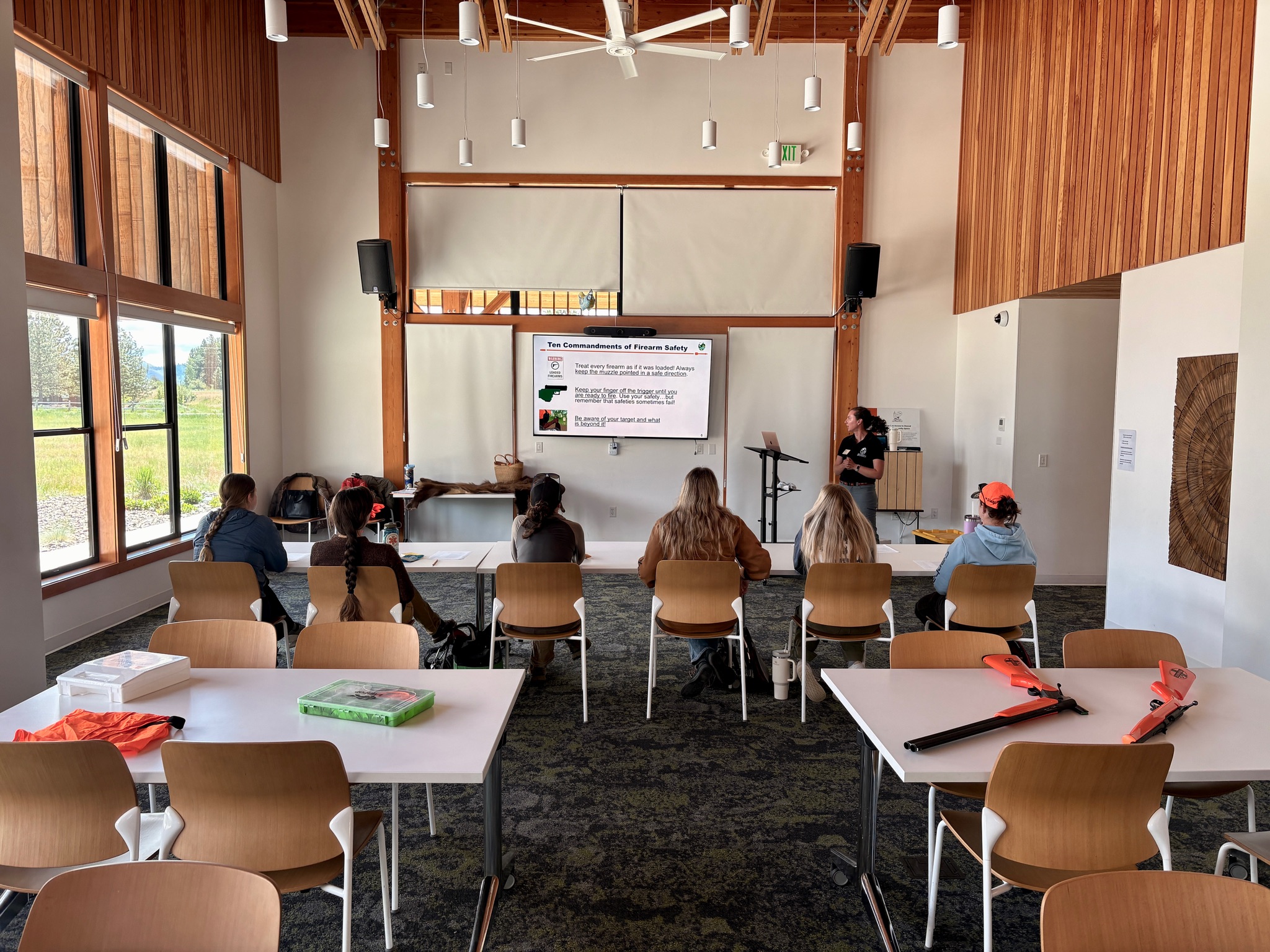 Women seated at tables in a high-ceilinged classroom looking at a screen displaying the Ten Commandments of Firearm Safety. Practice firearms with orange tips on the tables.