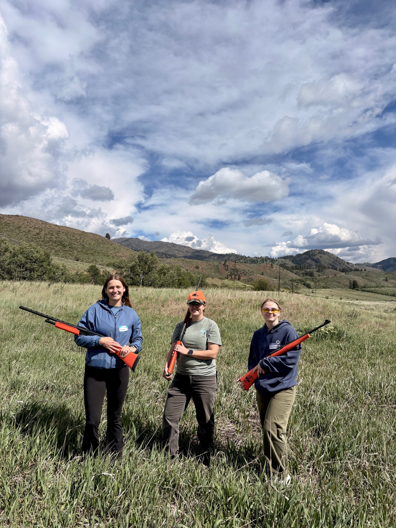 Three women holding practice firearms with orange tips, standing in a meadow with hills behind them under a Methow Valley sky.